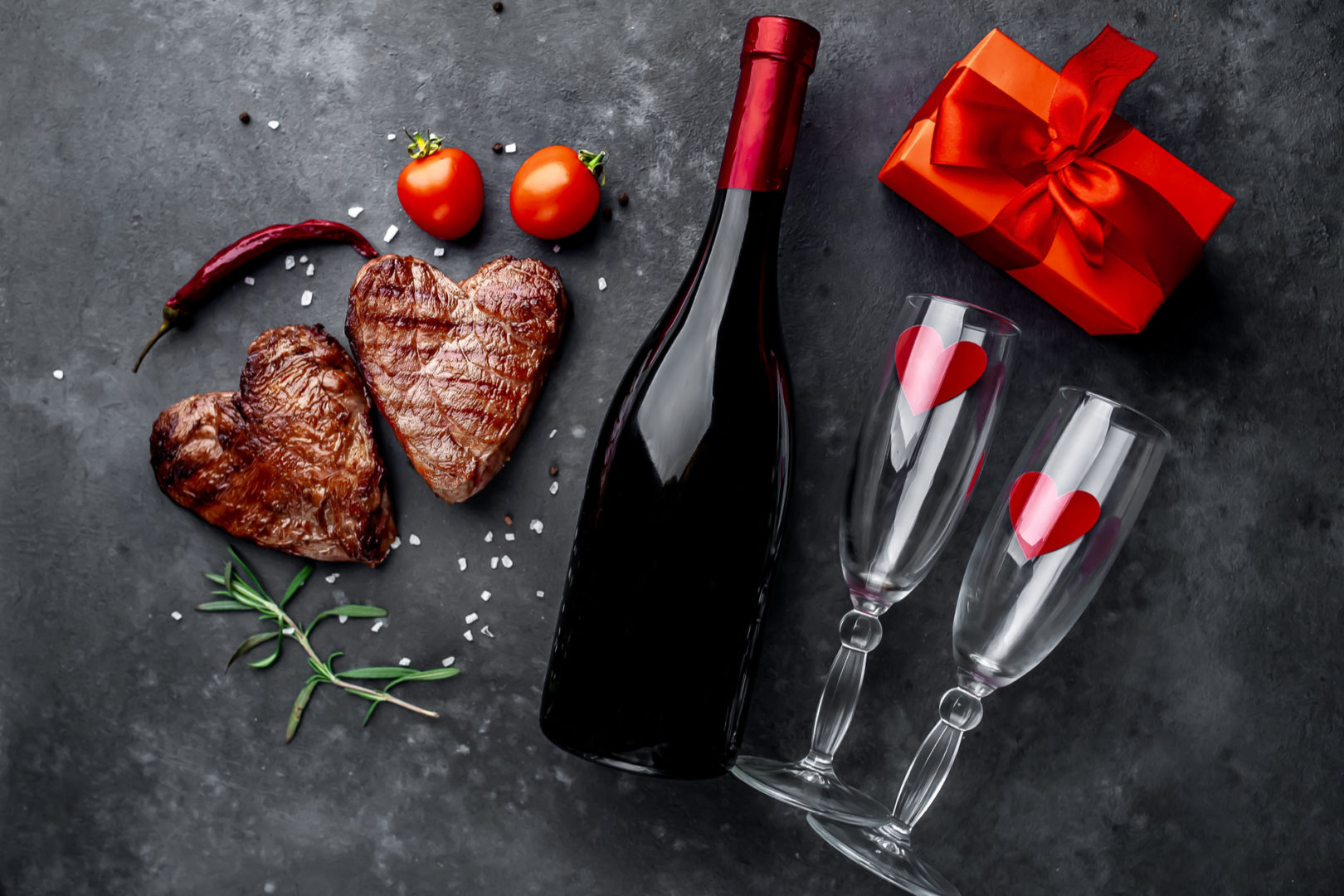 two grilled beef steaks in the form of a heart with spices, a gift with a red ribbon and a bottle of champagne or wine with two glasses for dinner for Valentine's day on a stone